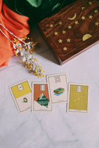 Four illustrated tarot cards arranged on a light marble-like surface beside an open wooden box decorated with gold stars and crescent moons, with orange fabric and dried yellow flowers nearby.