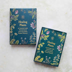Two boxed card decks titled "Healing Plants" arranged on a light stone surface. The boxes are dark teal with gold botanical illustrations and white text, and one box is slightly open to show the card arrangement inside.