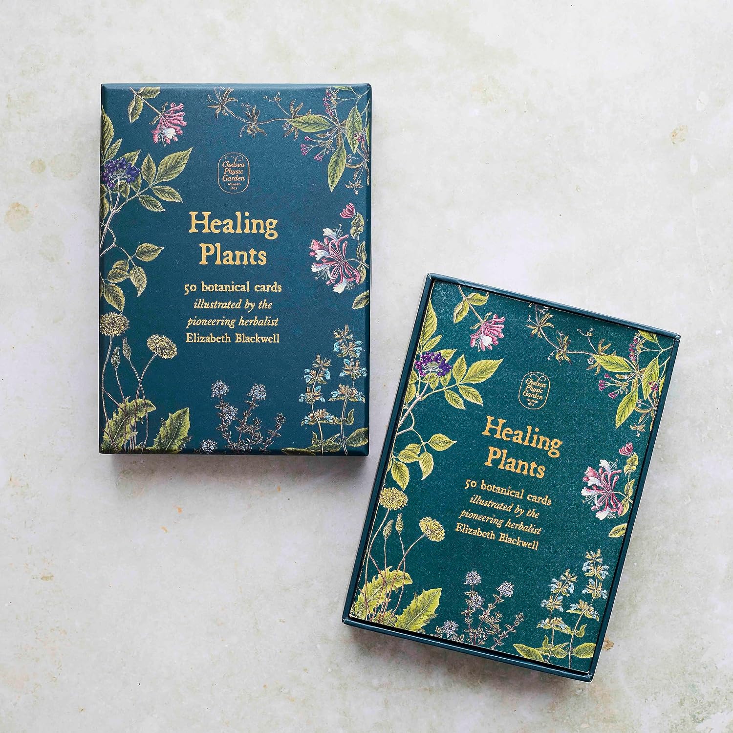 Two boxed card decks titled "Healing Plants" arranged on a light stone surface. The boxes are dark teal with gold botanical illustrations and white text, and one box is slightly open to show the card arrangement inside.