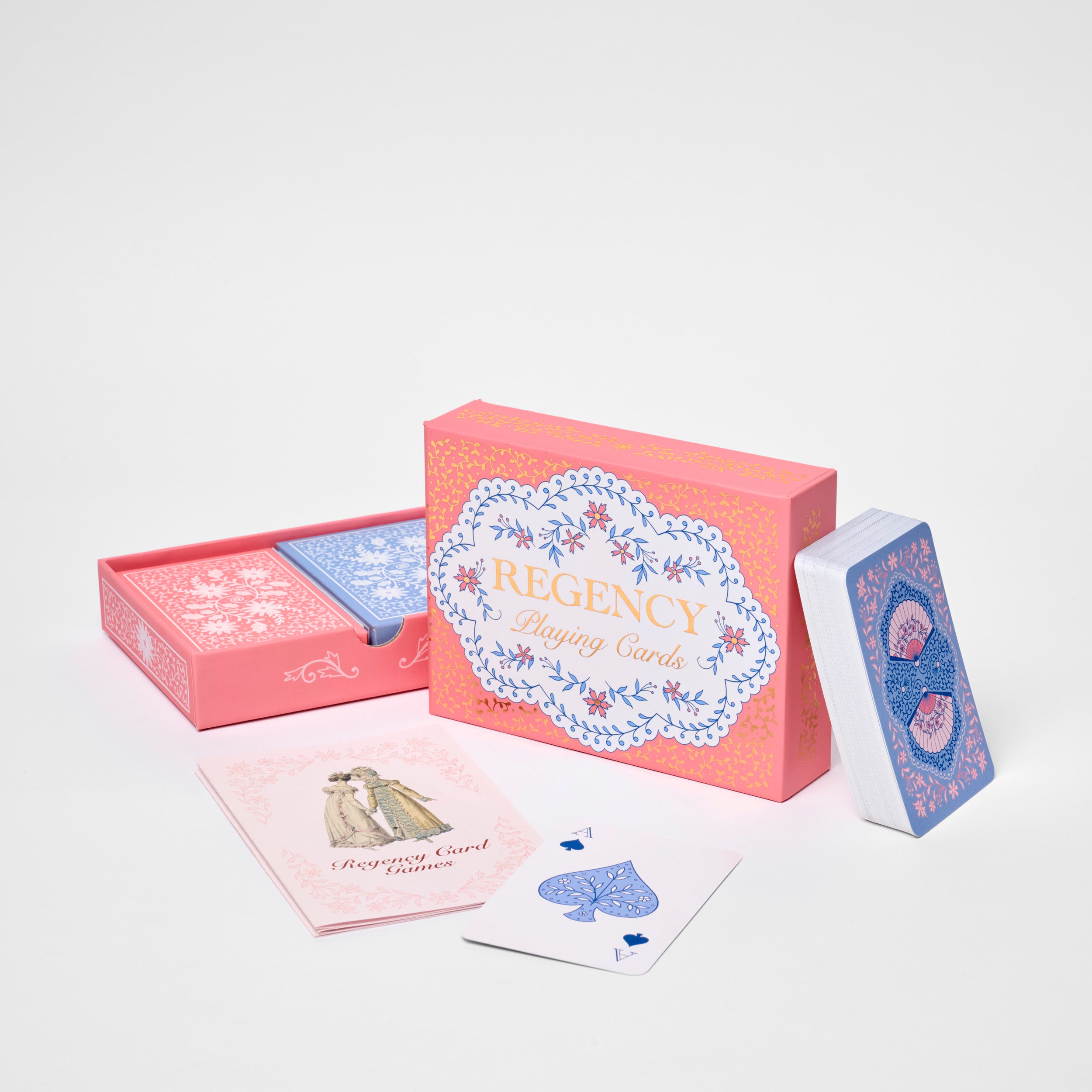 A boxed deck of playing cards called REGENCY, shown with its coral-pink box, an open pink drawer-style box, a blue-backed card deck standing upright, and two sample cards in front on a clean white studio background.