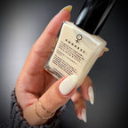 A hand with long, glossy off-white manicured nails holds a square glass bottle of Goddess nail polish. The bottle contains a creamy off-white polish and features a black cap, with the brand name 'GODDESS' printed on the front.