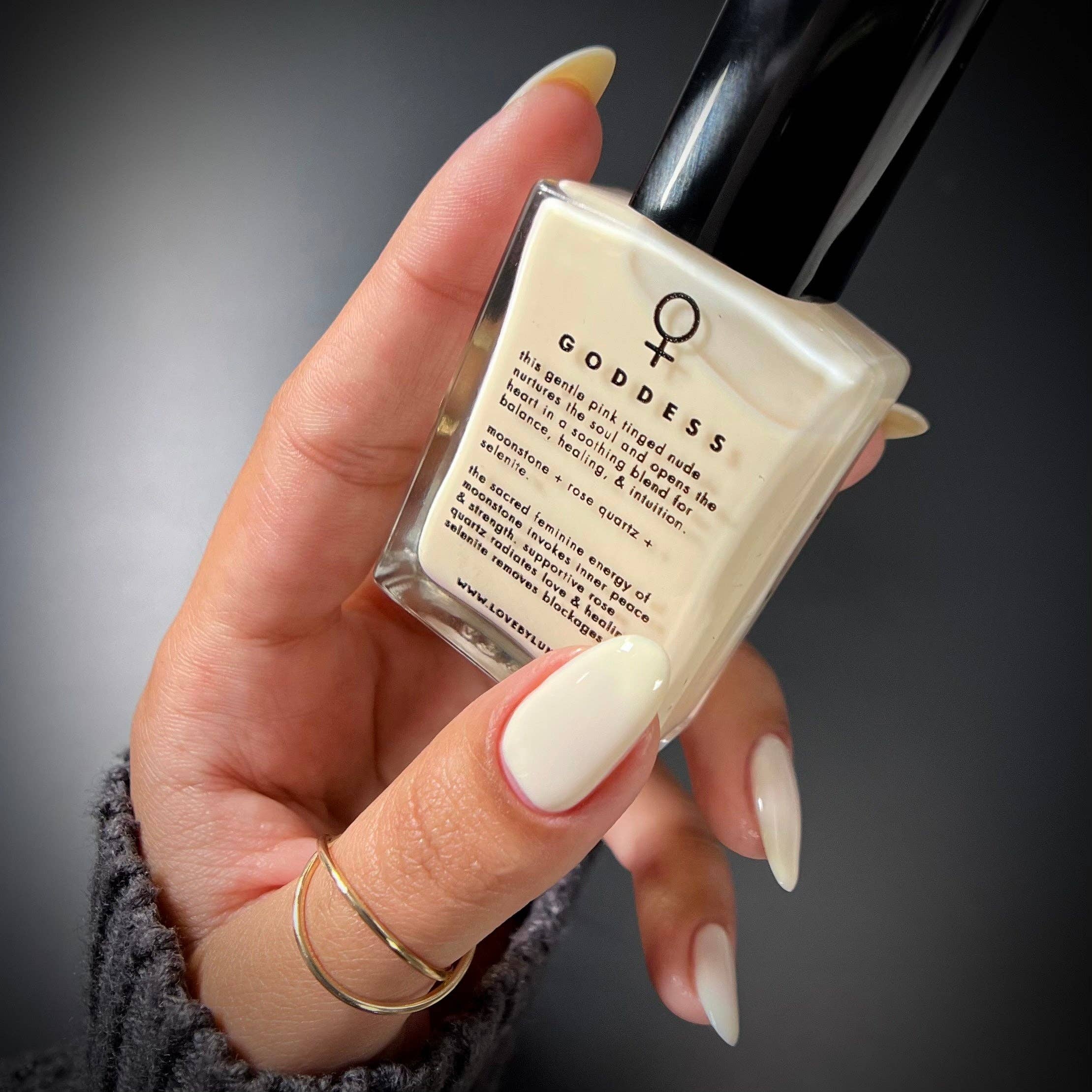 A hand with long, glossy off-white manicured nails holds a square glass bottle of Goddess nail polish. The bottle contains a creamy off-white polish and features a black cap, with the brand name 'GODDESS' printed on the front.