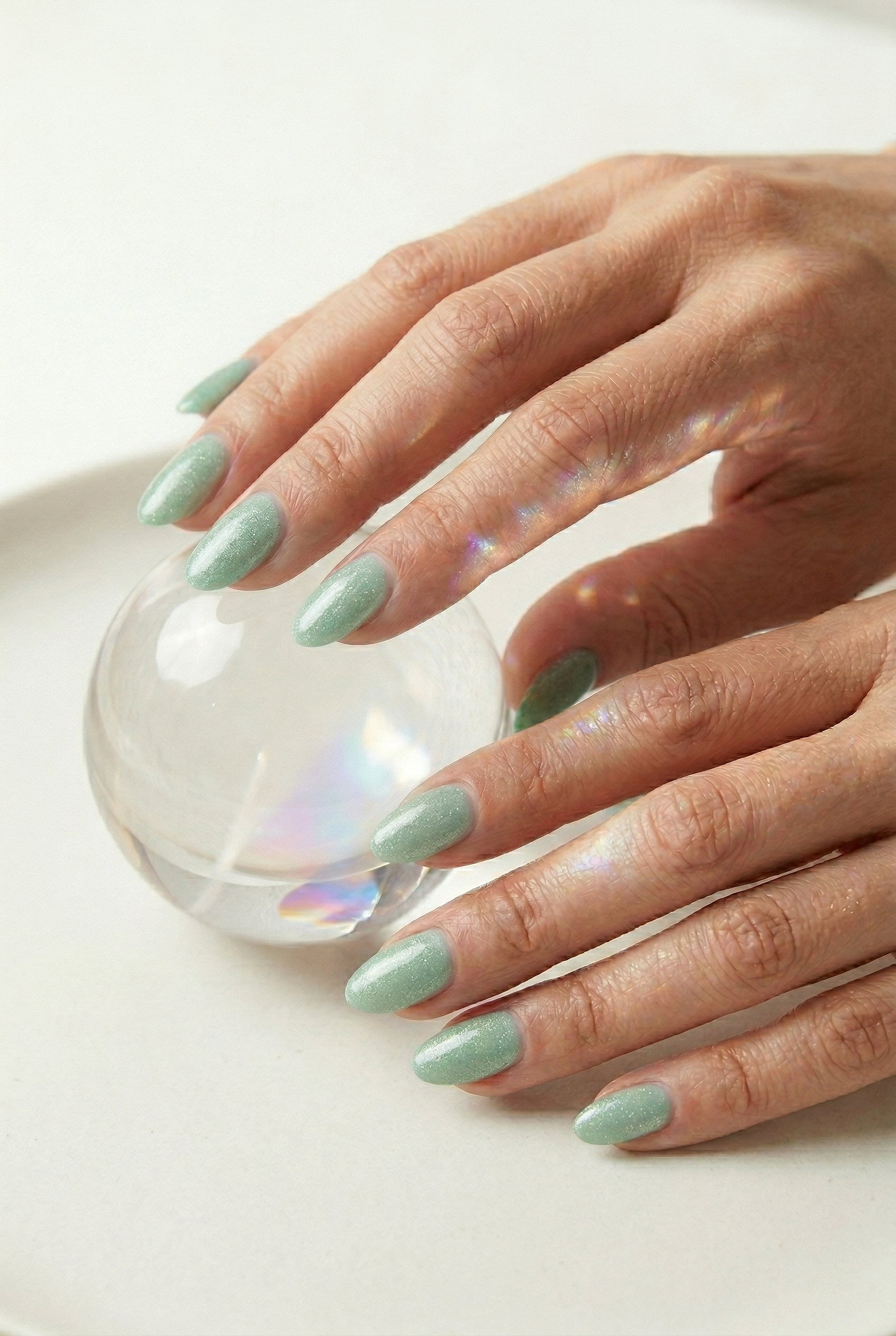Hand with light green nail polish holding a clear glass ball on a white background