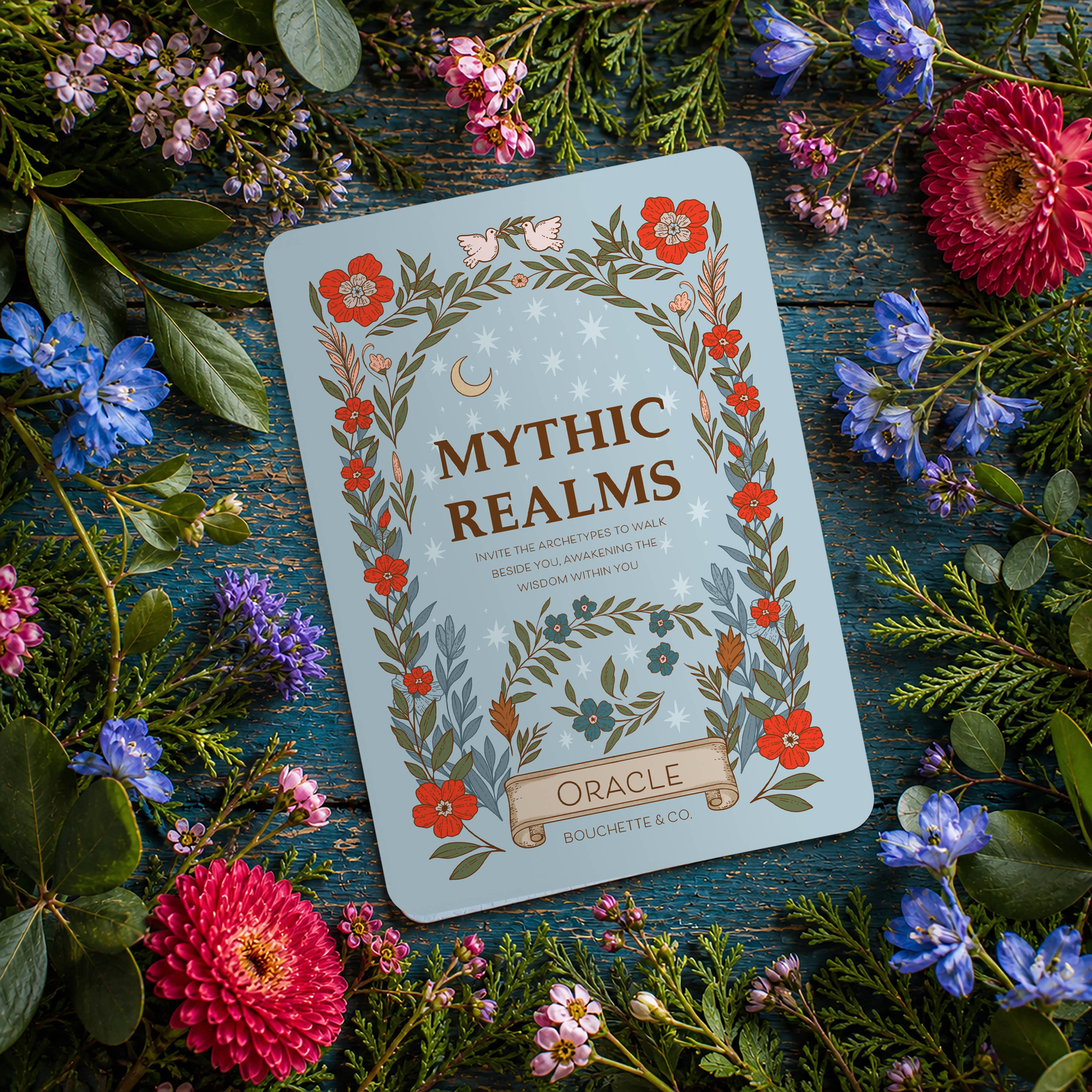 Ornate card titled 'Mythic Realms Oracle' surrounded by flowers on a wooden surface