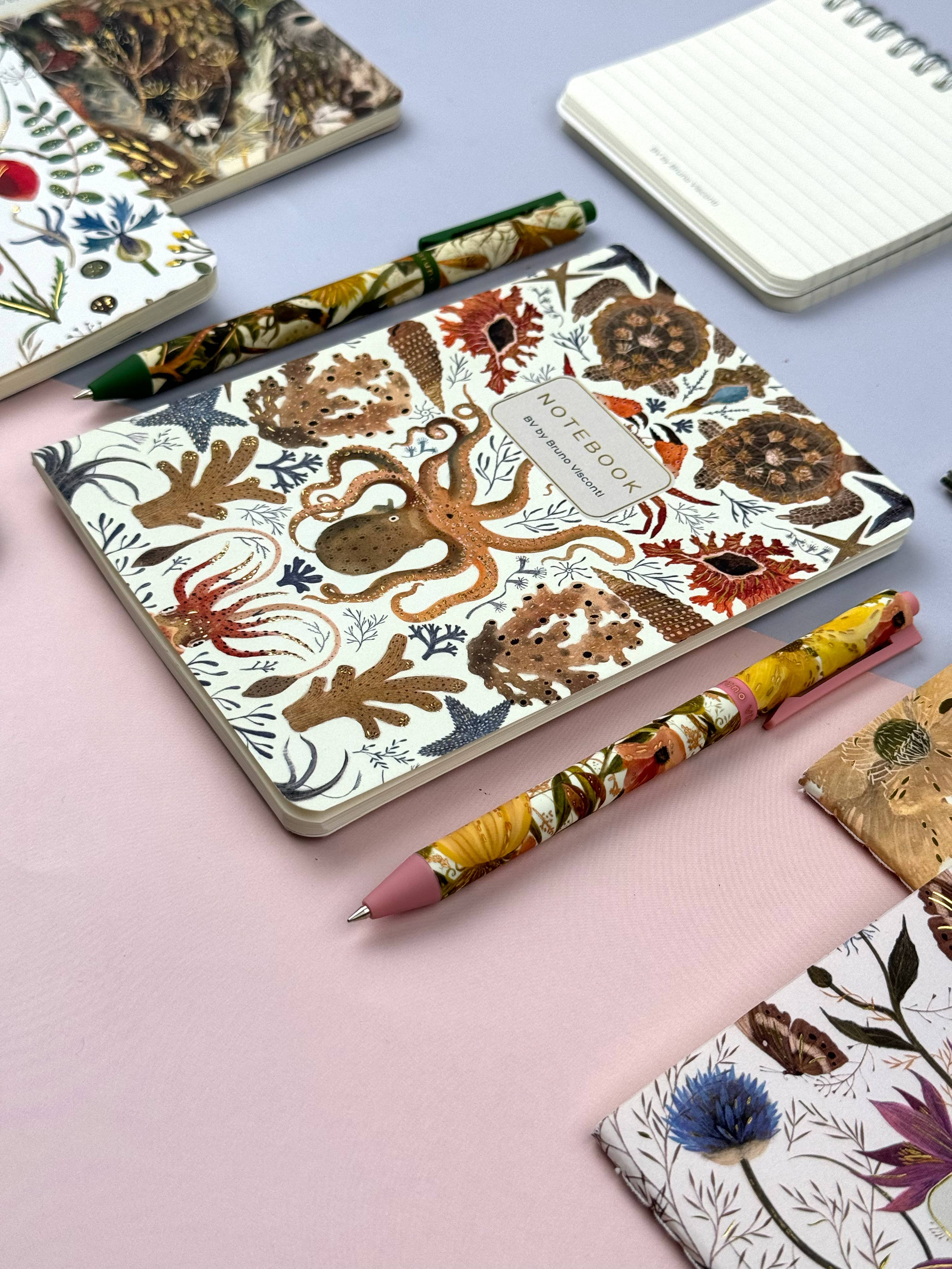 Notebooks and pens with nature-themed designs on a light pink surface