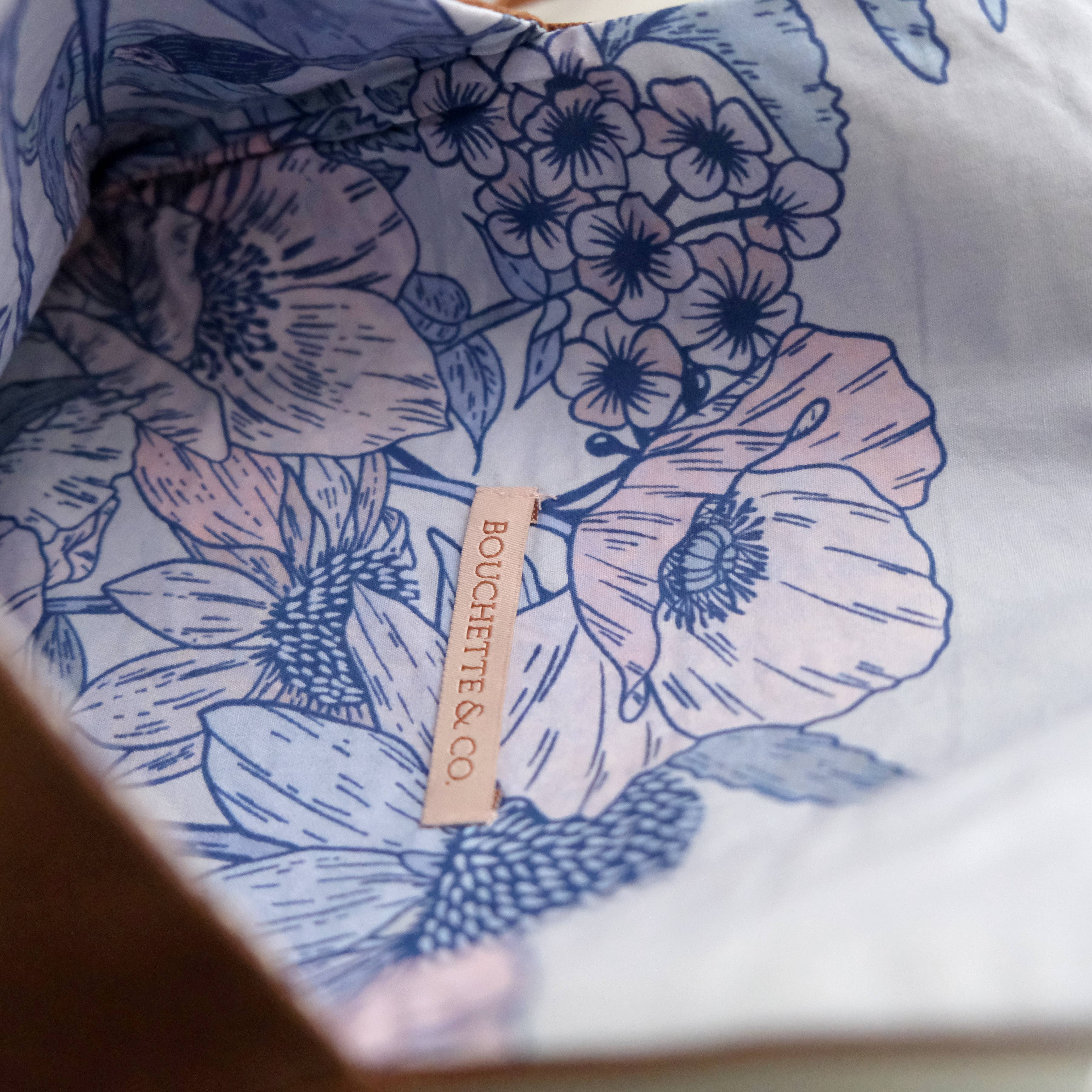 A close-up of a white fabric item, likely a scarf or bandana, featuring a blue and pink floral print. A beige paper brand tag reading 'BOUCHETTE & CO.