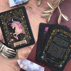 A styled flat-lay of a tarot deck featuring a black background with a pink unicorn illustration on the top card labeled 'UNICORN,' and several cards fanned out showing oracle text under the name 'HECATE.' The scene is arranged on a wooden surface with decorative crystals, a metallic hand-shaped object, and a gold branch.