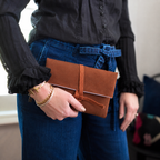 Person holding a brown leather clutch with a strap, wearing a black shirt and blue jeans.