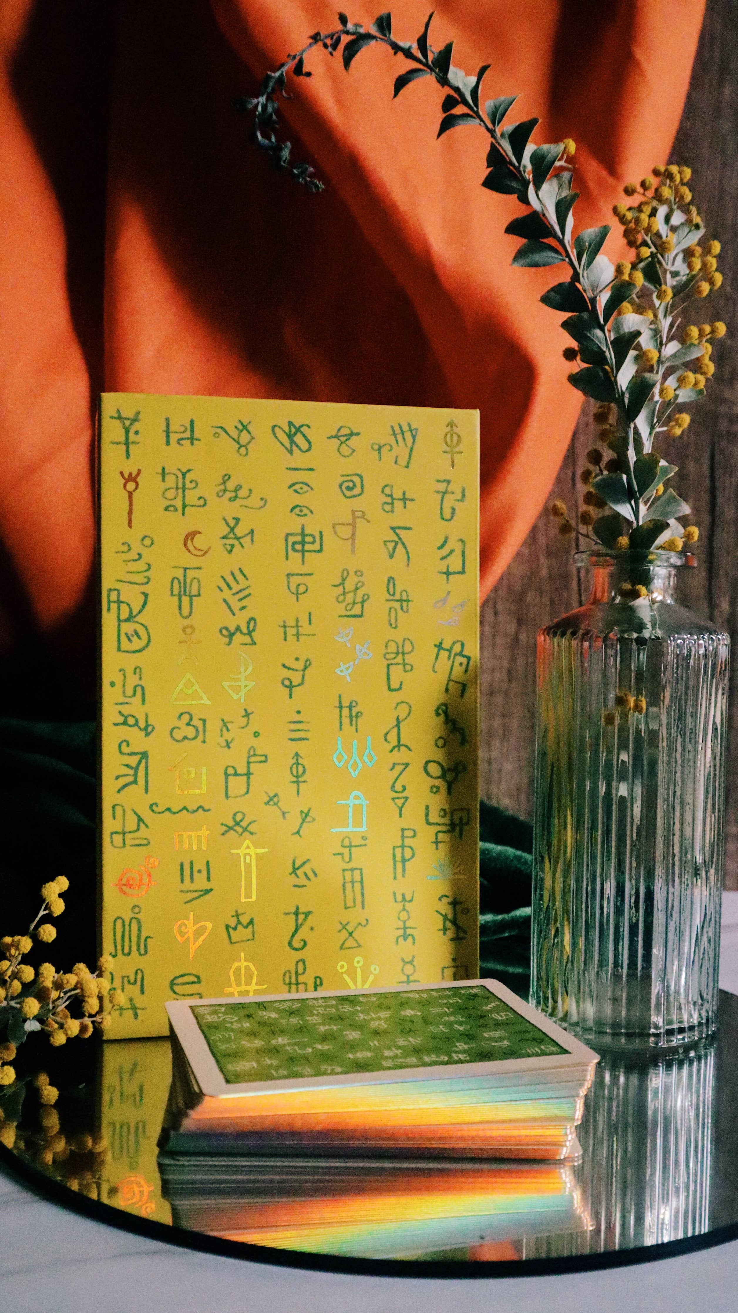 A yellow oracle card deck with green and gold ancient script symbols, shown with a stack of cards on a reflective surface in front of a draped orange fabric backdrop and a glass vase with greenery.