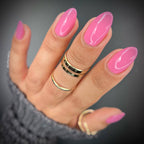 A close-up of a hand with glossy bright pink almond-shaped nails, wearing multiple gold rings and a gray knit sweater cuff, against a dark gray background.