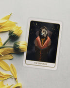 A tarot card titled 'THE EMPEROR' lies on a light gray surface next to yellow flower buds. The card features an illustration of a dog dressed in a dark robe with a red-orange collar against a starry night sky.