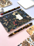 Floral notebook and pen on a light pink surface with other stationery items.