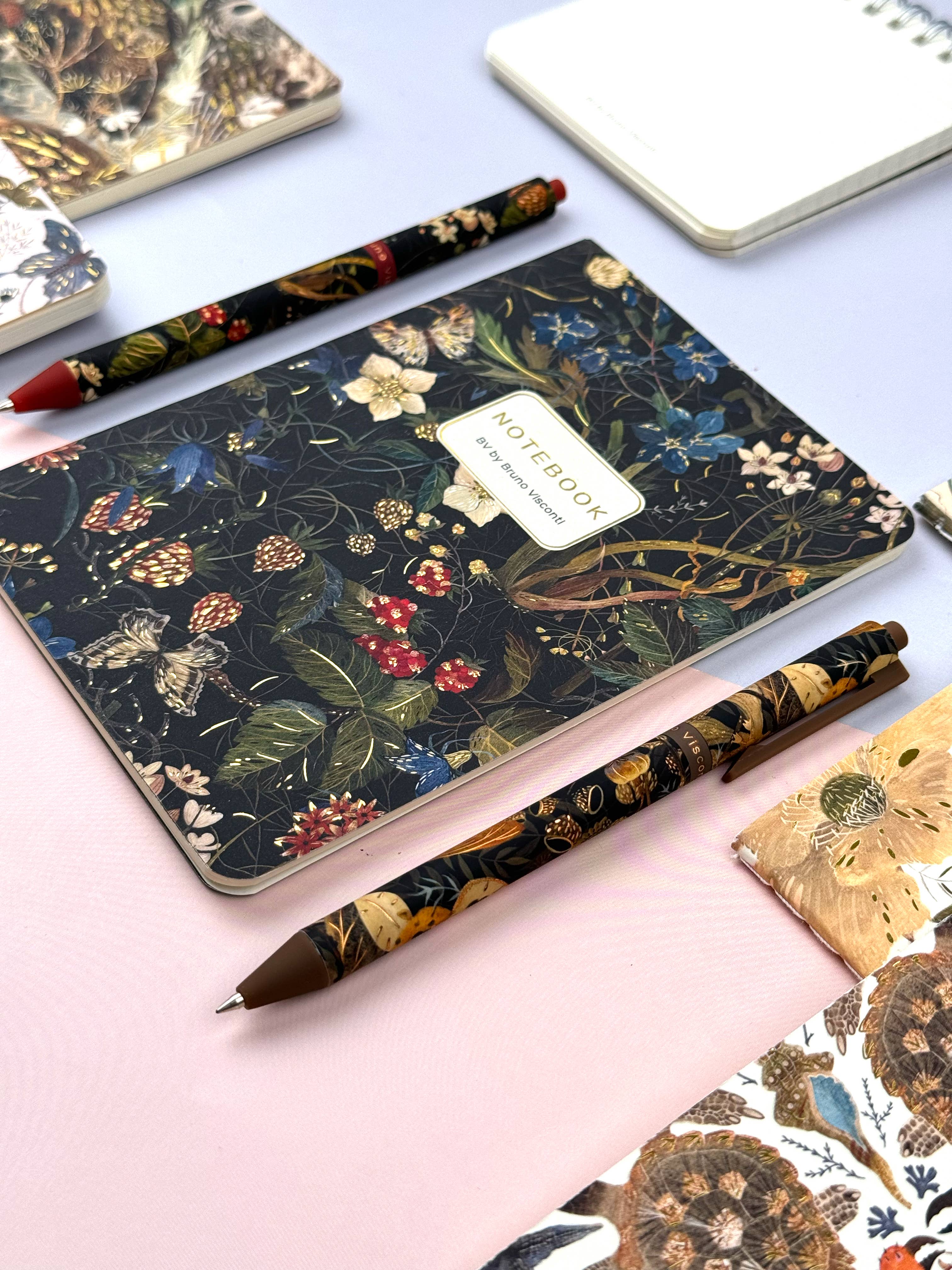 Floral notebook and pen on a light pink surface with other stationery items.