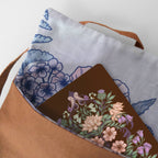 A close-up of a brown canvas tote bag with a white and blue floral patterned lining, holding a brown notebook decorated with a colorful floral and bird illustration.
