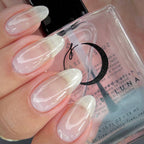 A close-up of a hand with long, glossy almond-shaped nails painted in a soft sheer pink and white gradient, holding a bottle of BY LUNA infused polish.