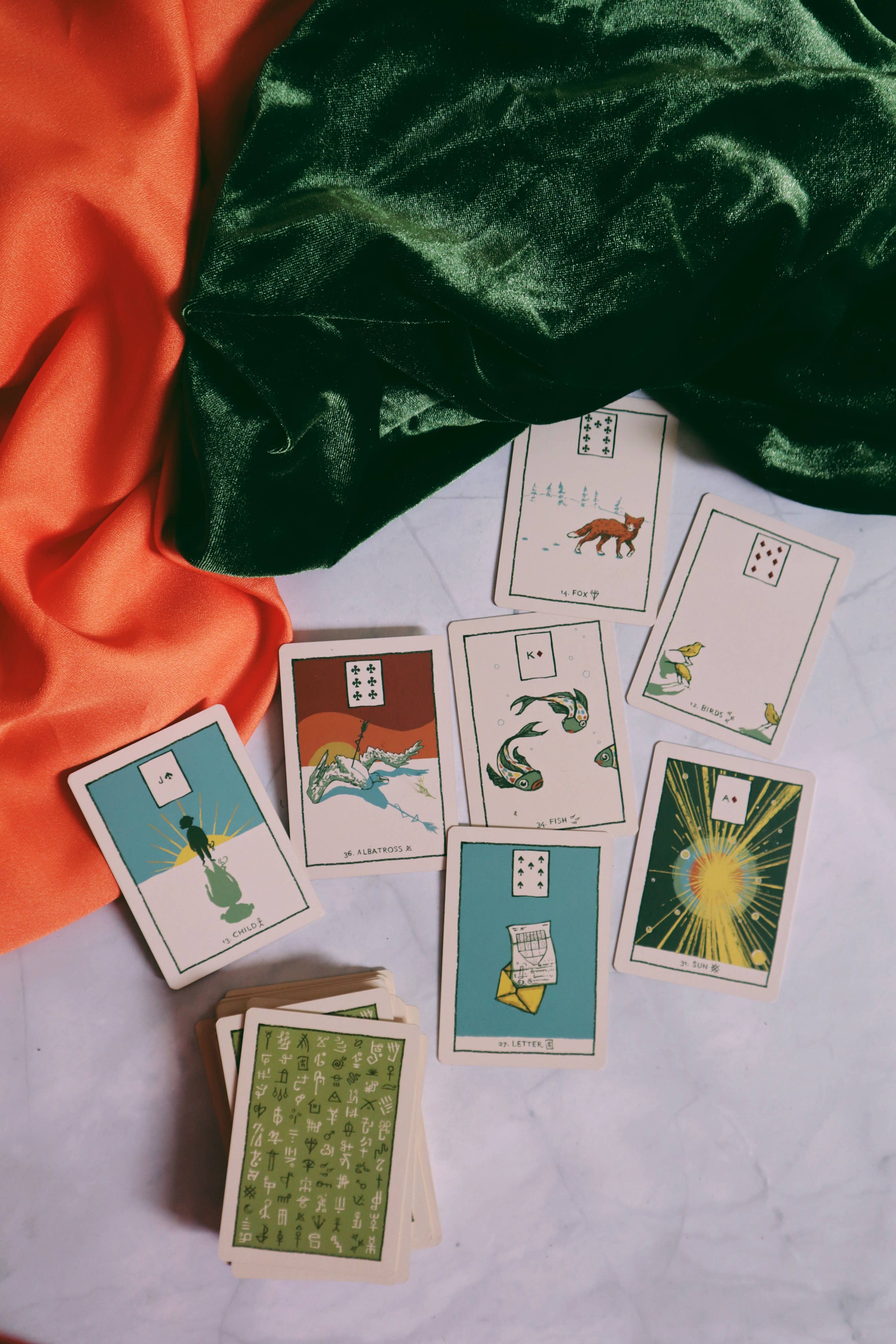A spread of illustrated tarot or oracle cards arranged on a light surface with draped orange and dark green fabric around them. The cards show simple artwork and numbers, with one card back visible showing a green patterned design.