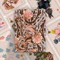 A rectangular notebook or journal with a vintage botanical pattern featuring pink flowers and brown leaves on a light beige background, styled on top of a collage of illustrated playing cards.