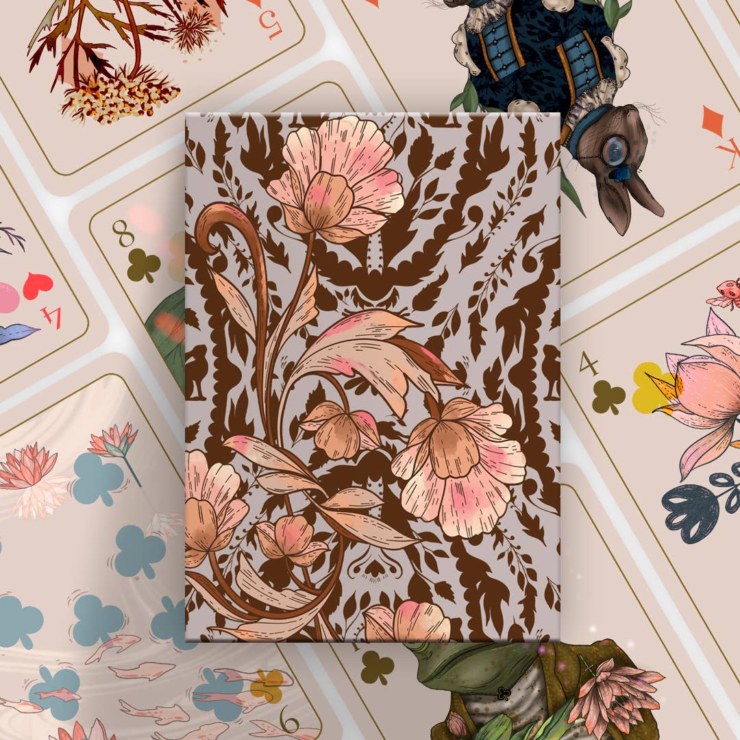 A rectangular notebook or journal with a vintage botanical pattern featuring pink flowers and brown leaves on a light beige background, styled on top of a collage of illustrated playing cards.
