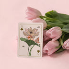 A single illustrated playing card with a lotus flower and club suit design stands upright beside a bouquet of pink tulips on a soft pink background.