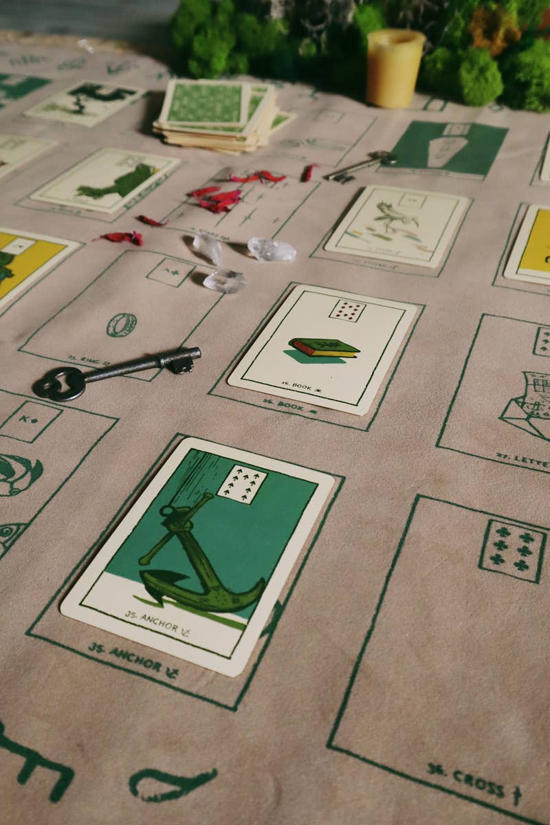 A close-up of a vintage-style card game board or playmat spread out on a table, featuring illustrated cards with anchors, letters, and symbols on a beige background. A small stack of cards and a candle are visible in the background.