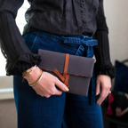 Person holding a dark gray clutch with a brown strap, wearing a black shirt and blue jeans.