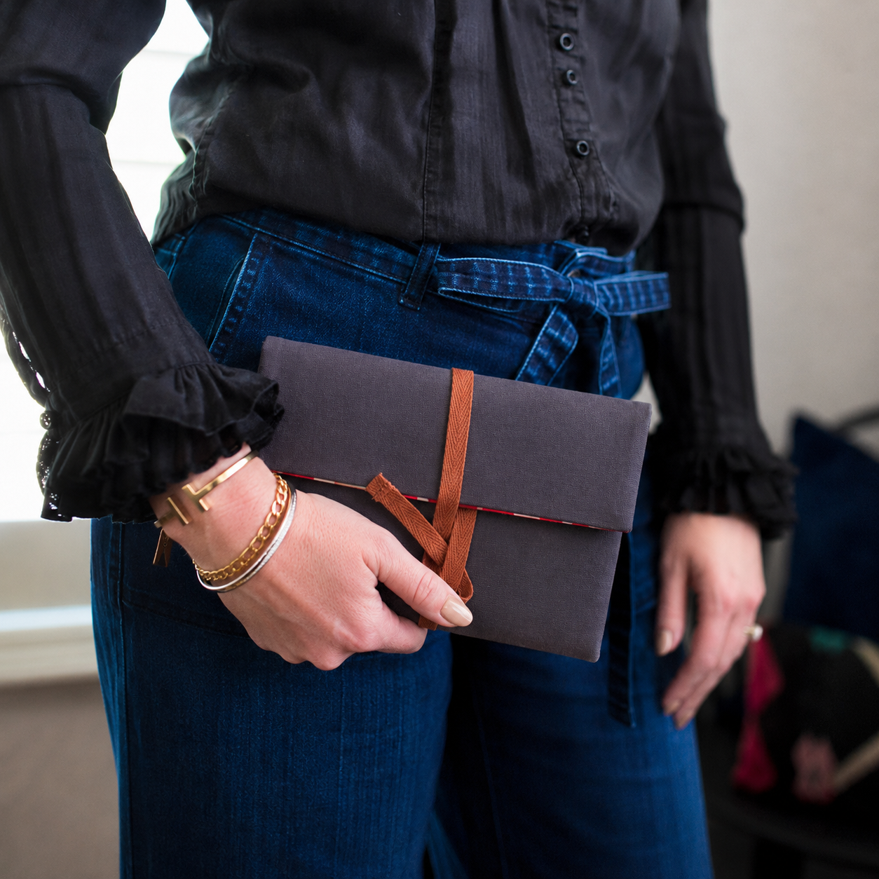 Person holding a dark gray clutch with a brown strap, wearing a black shirt and blue jeans.