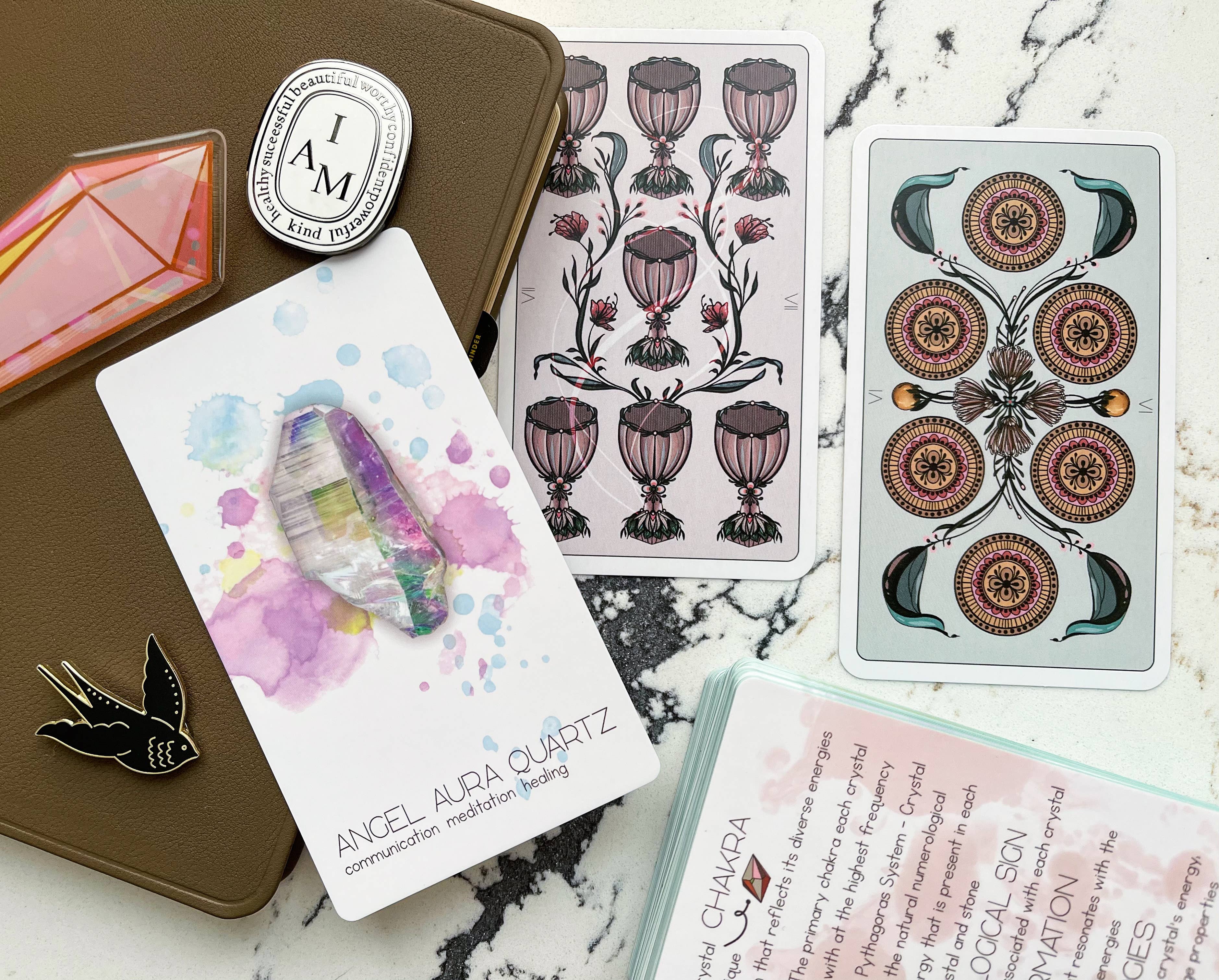 A styled flat-lay of a tarot card deck and oracle card set on a white marble surface, alongside a brown journal, a crystal, and a bird enamel pin.