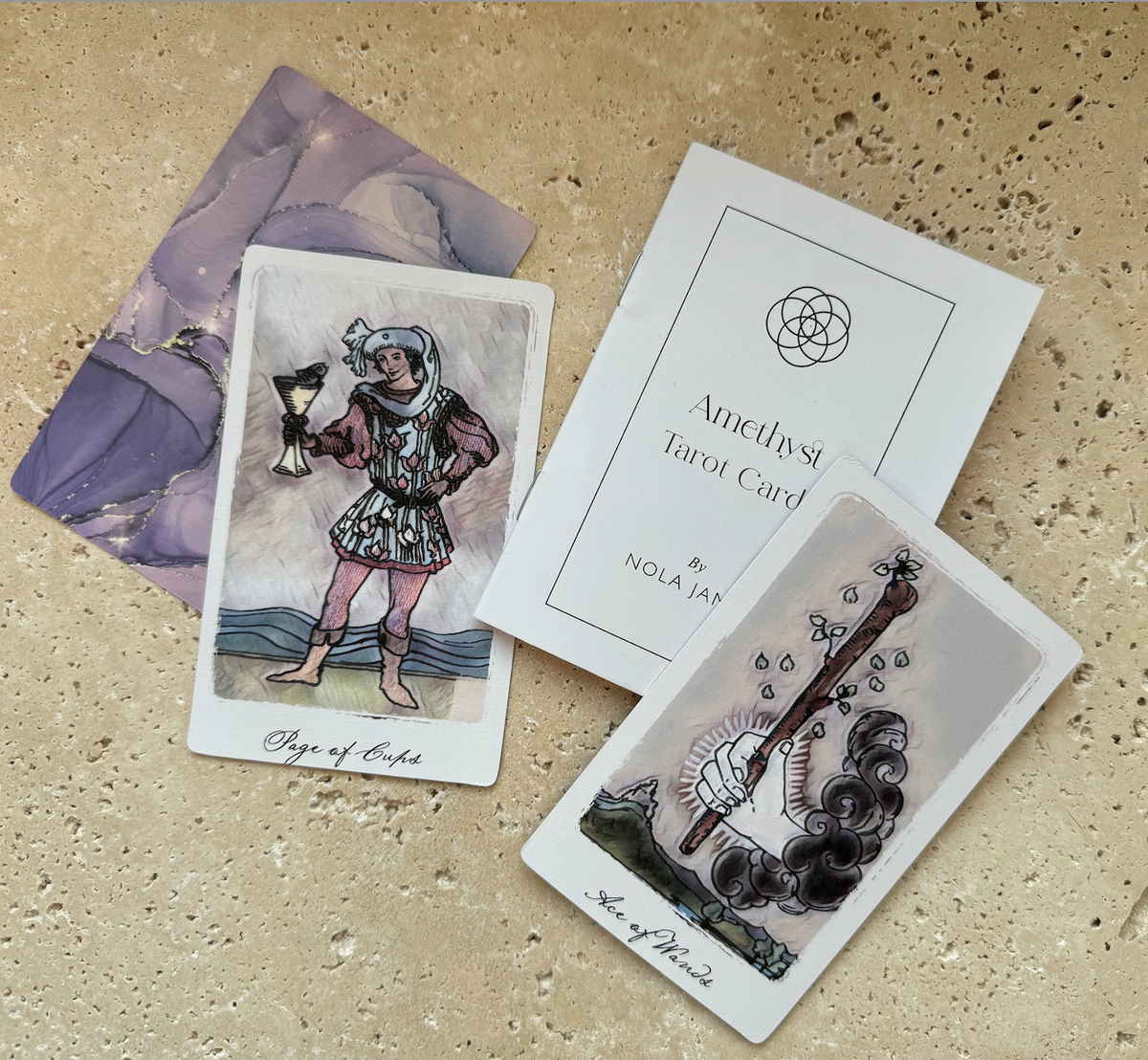 A tarot card deck laid out on a beige stone surface, showing the back of a purple card, a white card titled 'Amethyst Tarot Card' by Nola Jan, a 'Page of Cups' card, and a 'Page of Swords' card.