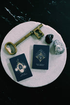 Two tarot cards with ornate gold and white designs on a dark navy background, arranged on a round white stone tray with a vintage gold key, a black crystal, and a white crystal on a black marble surface.