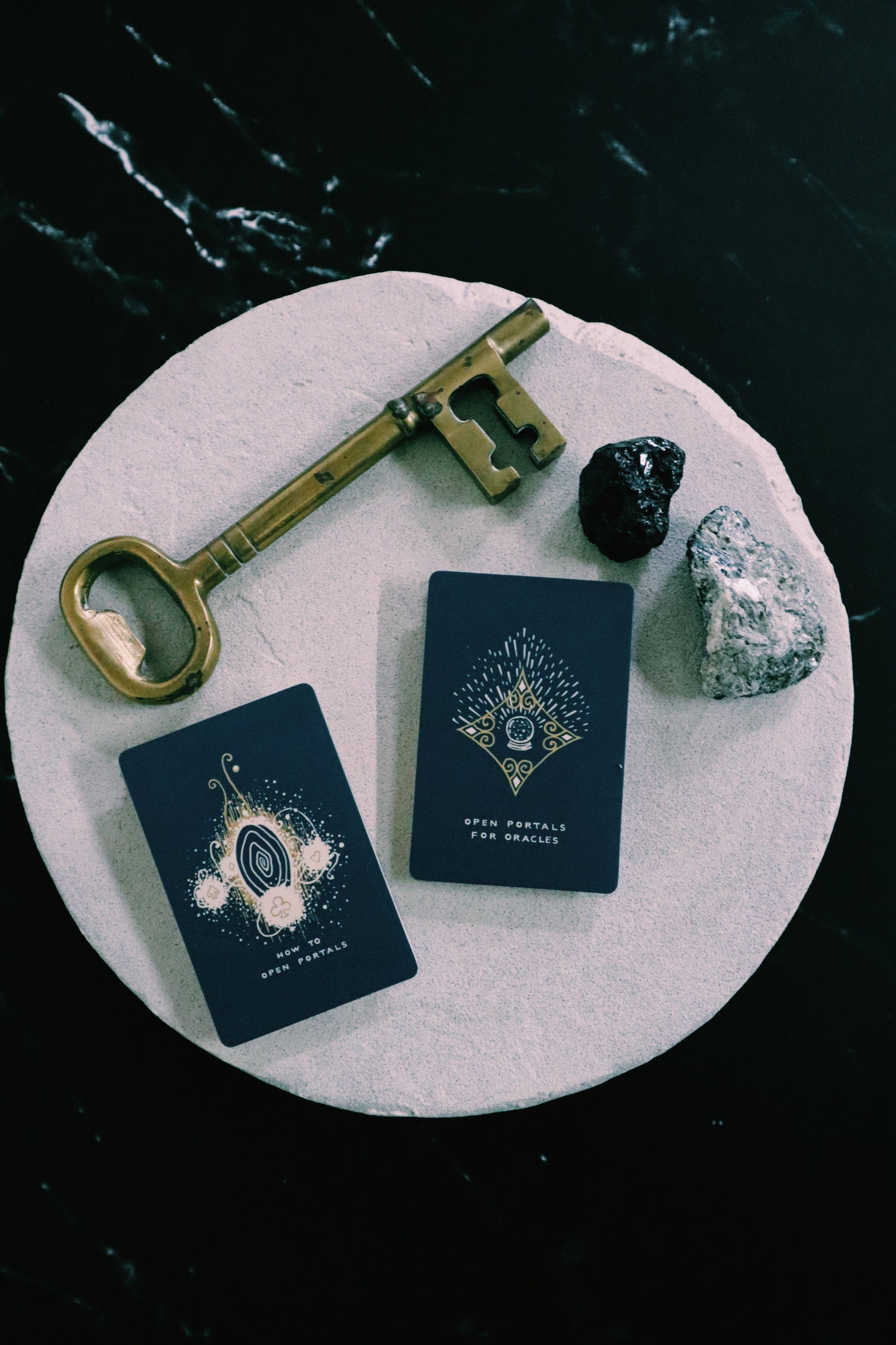 Two tarot cards with ornate gold and white designs on a dark navy background, arranged on a round white stone tray with a vintage gold key, a black crystal, and a white crystal on a black marble surface.