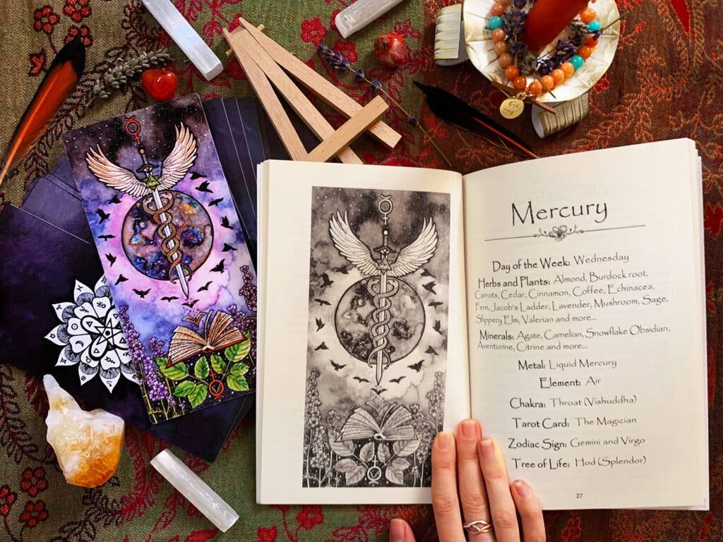 An open book displaying a black-and-white illustration of Mercury with wings and a caduceus symbol on one page, and the Day of the Week entry for Wednesday on the other. The scene is styled as a flat lay with tarot cards, crystals, a feather quill, beads, and other mystical props on a patterned red and green cloth background.