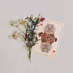 A decorative playing card featuring an illustration of a fluffy gray cat in a brown kimono with red hearts, styled with artificial white flowers and green stems on a light gray background.
