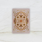 A rectangular decorative card or tile with rounded corners, featuring an ornate symmetrical floral and scroll pattern in beige, brown, and muted pink tones, placed on a light marble-like surface.