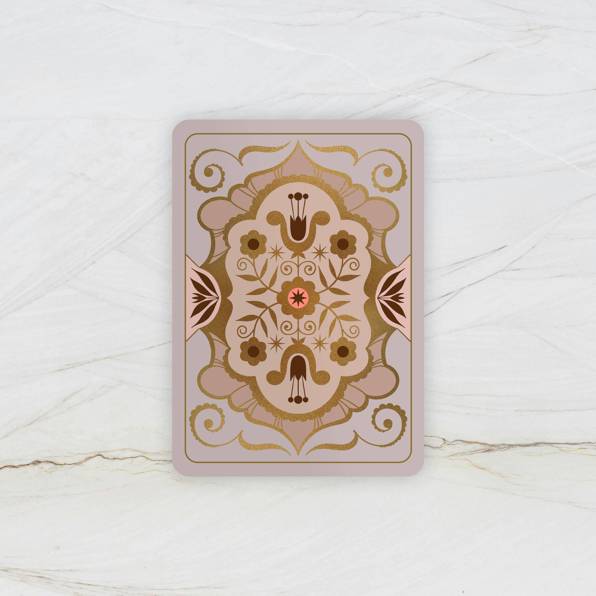 A rectangular decorative card or tile with rounded corners, featuring an ornate symmetrical floral and scroll pattern in beige, brown, and muted pink tones, placed on a light marble-like surface.