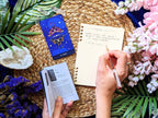 Person reading a tarot card with a notebook and pen on a woven mat surrounded by flowers and plants.