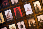 A close-up of multiple tarot or oracle cards laid out on a wooden surface. The cards have dark borders and show different illustrated and photographic portraits, including a central card with a red-tinted dark figure and other cards with sepia-toned figures.