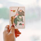 A hand with glittery red nail polish holds a playing card featuring a whimsical illustration of leaves and hearts. The card is shown against a soft, blurred background with a hint of a window or light source.