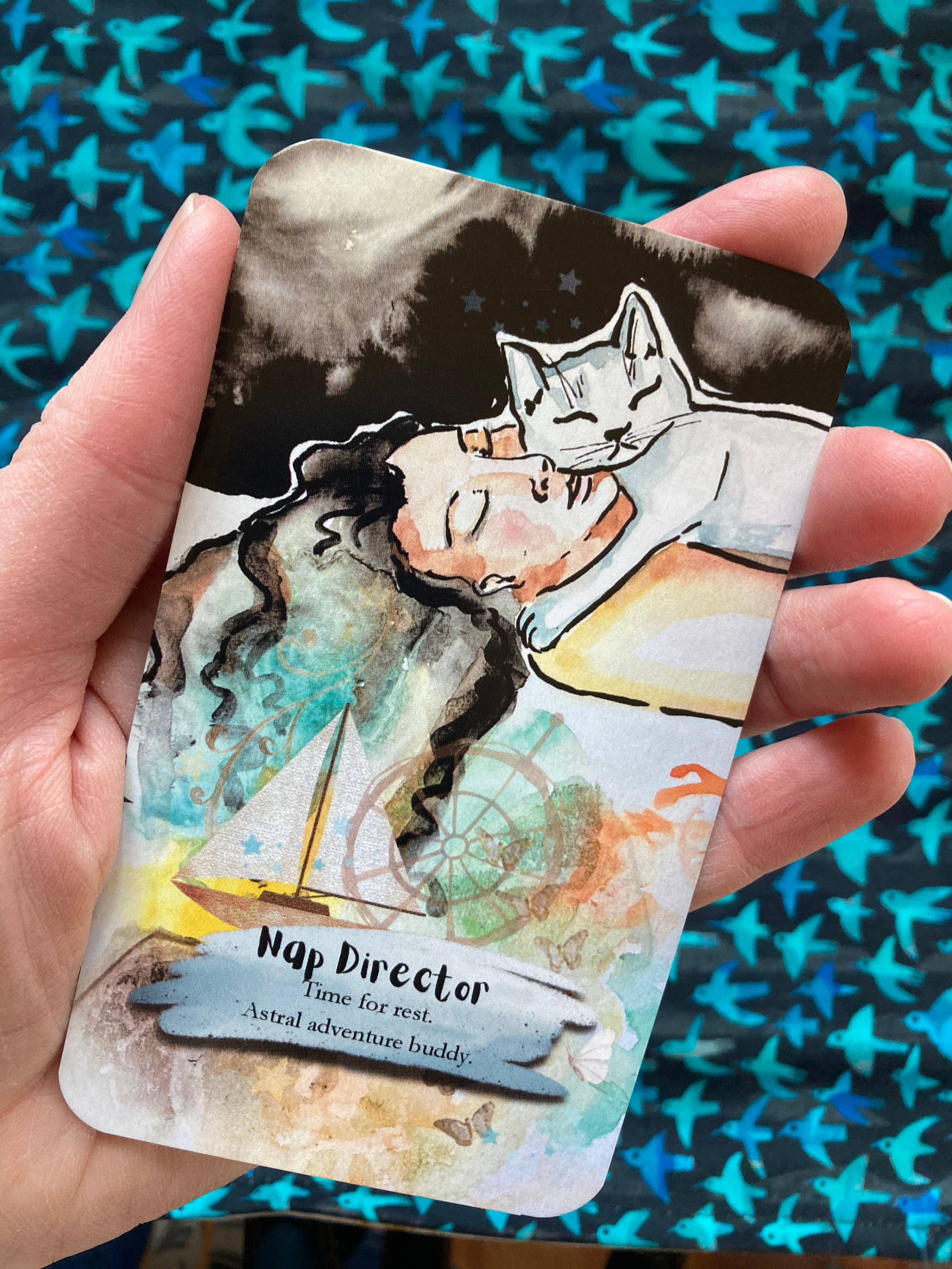 A hand holds a watercolor-style card featuring a sleeping woman and a white cat, with the text 'Nap Director' and 'Time for rest, Astral adventure buddy.' The background is a blue patterned fabric.