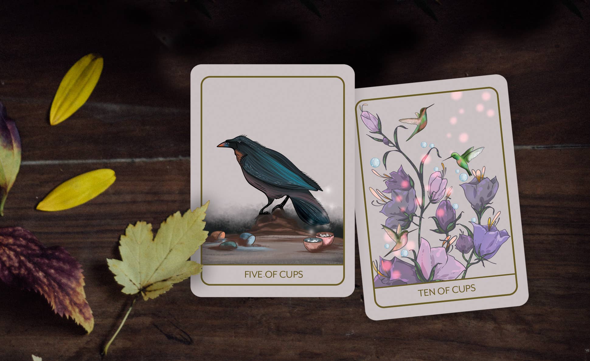 Two illustrated tarot cards, labeled 'FIVE OF CUPS' and 'TEN OF CUPS,' placed on a dark wooden surface with scattered autumn leaves. The left card shows a blue-black bird, and the right card shows purple flowers and small hummingbirds.