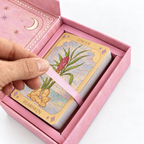 A hand is holding a pink ribbon loop over a deck of tarot cards inside a pink gift box. The top card is labeled 'GINGER' and 'REAWAKEN', with botanical illustrations of ginger roots and leaves on a light blue and yellow background.