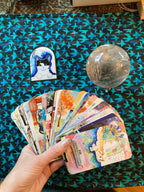A hand holds a fanned-out deck of colorful illustrated oracle or tarot cards over a dark patterned surface. A small illustrated card or magnet with a blue cat portrait and a clear glass orb sit nearby on the surface.