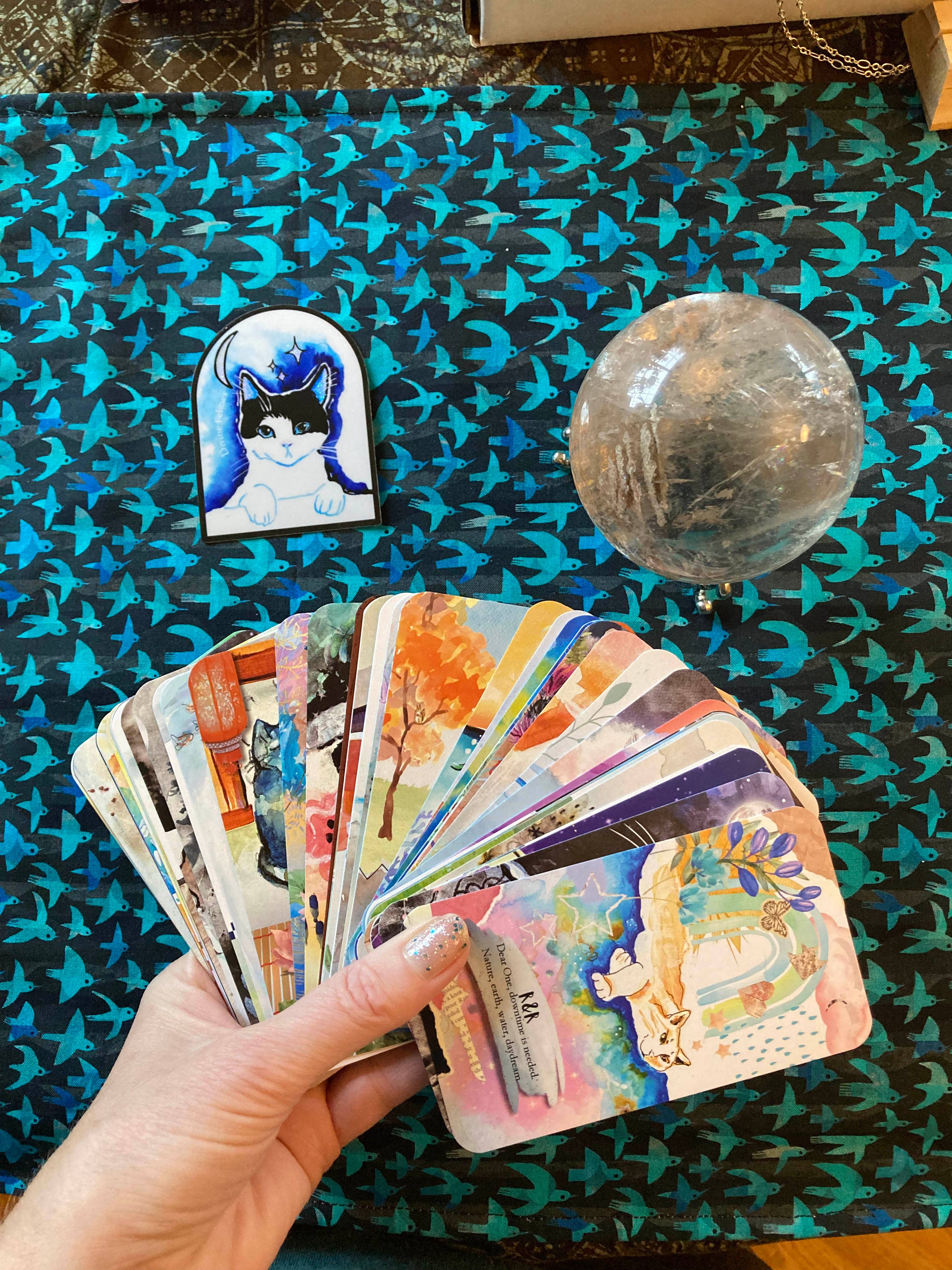 A hand holds a fanned-out deck of colorful illustrated oracle or tarot cards over a dark patterned surface. A small illustrated card or magnet with a blue cat portrait and a clear glass orb sit nearby on the surface.