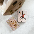 A deck of illustrated playing cards on a light textured surface, with one card face-up showing a bird illustration and the back of another card visible below it. A decorative wooden object is partially visible at the top left.