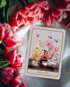 A tarot card reading 'SIX OF CUPS' is displayed on a light marble surface, surrounded by red and white tulips. The card features an illustration of flowers, bees, and candles.