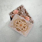 A square decorative card or tile with a gold and pink floral medallion design, partially sliding out of a matching patterned box on a light textured surface.
