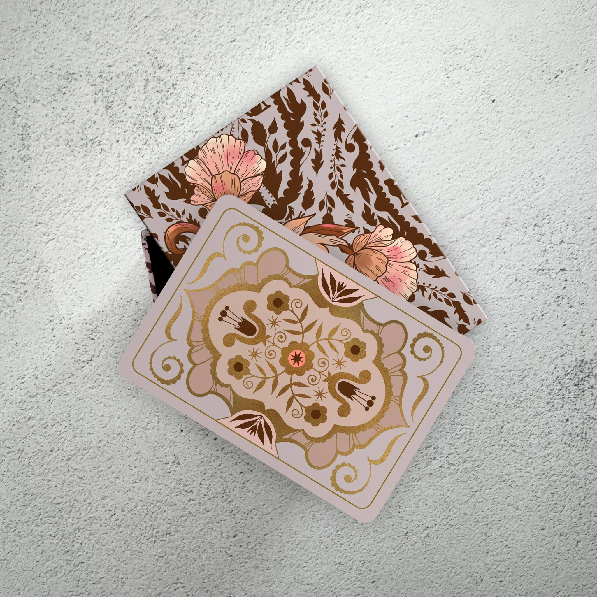 A square decorative card or tile with a gold and pink floral medallion design, partially sliding out of a matching patterned box on a light textured surface.