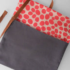 A tote bag with a dark gray front pocket, brown shoulder straps, and a top flap featuring a red polka dot pattern on a light beige background.