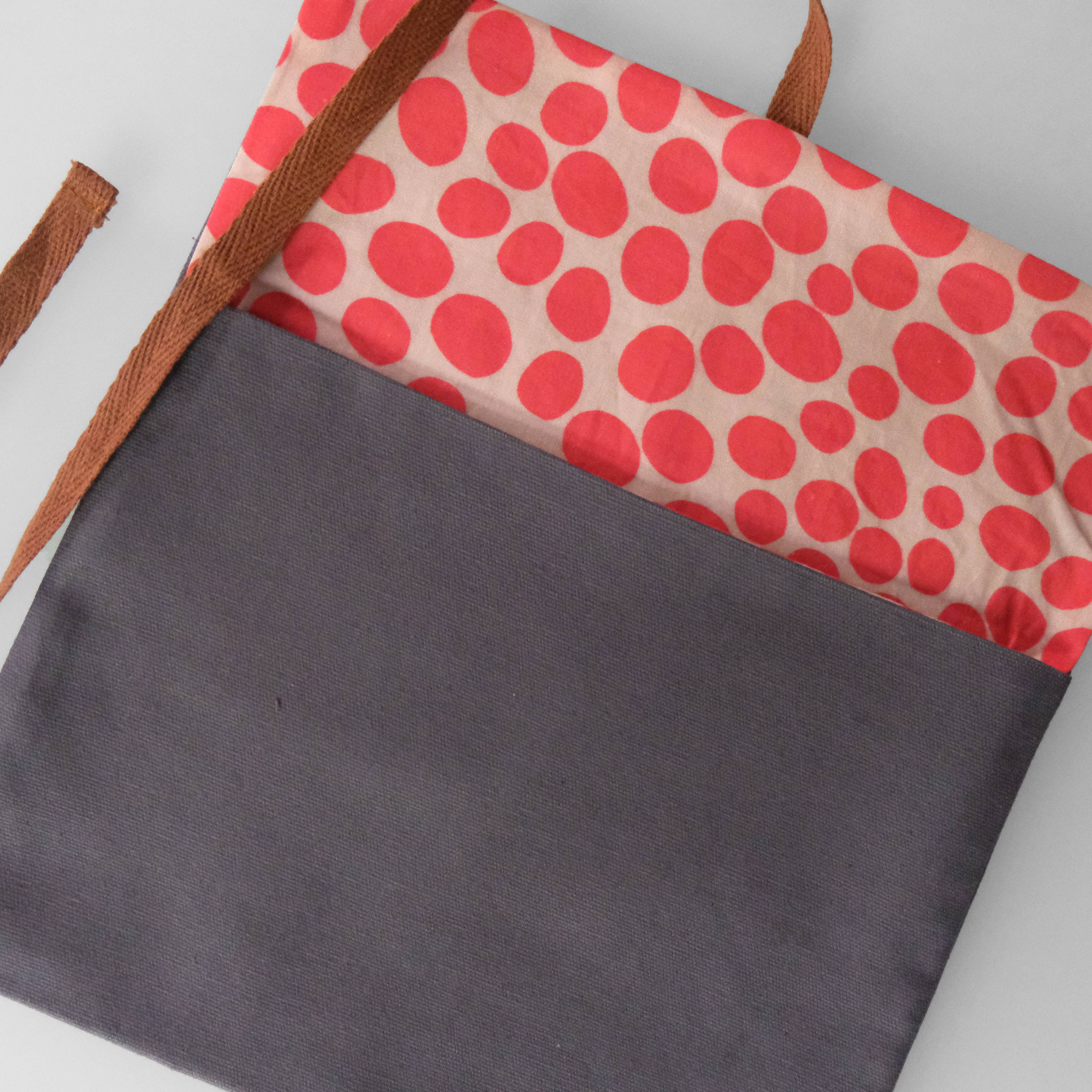 A tote bag with a dark gray front pocket, brown shoulder straps, and a top flap featuring a red polka dot pattern on a light beige background.