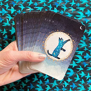 A hand holds a fanned deck of cards against a dark teal patterned background. The top card features an illustration of a blue cat inside a circular frame on a starry night sky background.