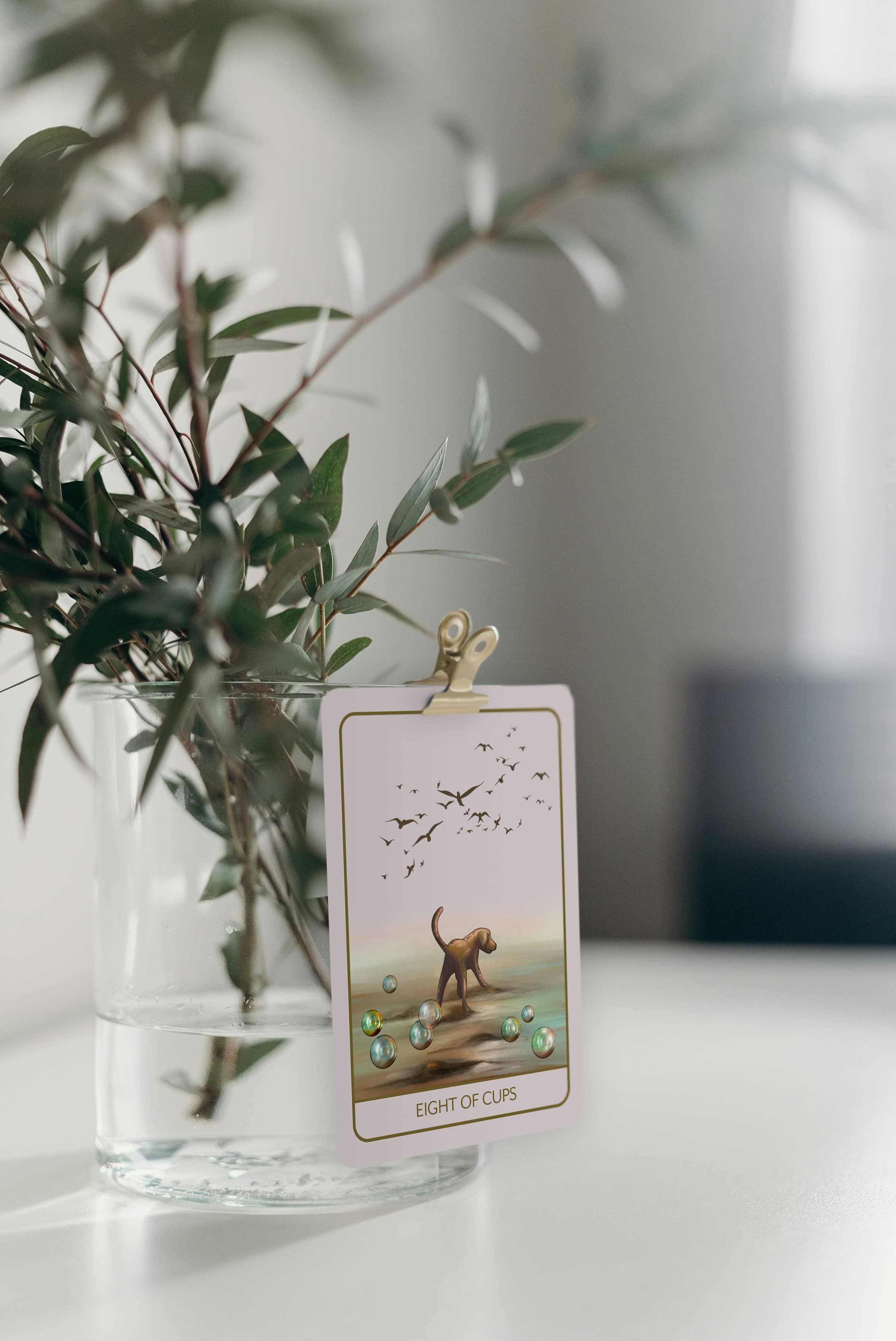 A tarot card, specifically the 'Eight of Cups' card, clipped to a gold metal clip and hanging from a clear glass vase filled with green leaves, set on a white tabletop.